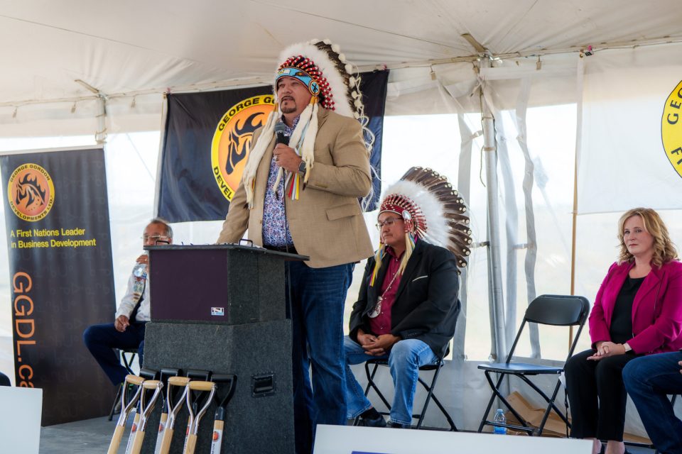 Des responsables de la PNGG, de SaskPower et de la mine de potasse K+S lors de la cérémonie d’annonce de Wicehtowak sous une tente blanche; un orateur se tient à un podium, trois hommes coiffés de coiffes à plumes et deux autres sont assis derrière l’orateur; des pelles cérémonielles sont alignées devant l’orateur. Les drapeaux de la Première Nation George Gordon et de George Gordon Developments Ltd. sont visibles.