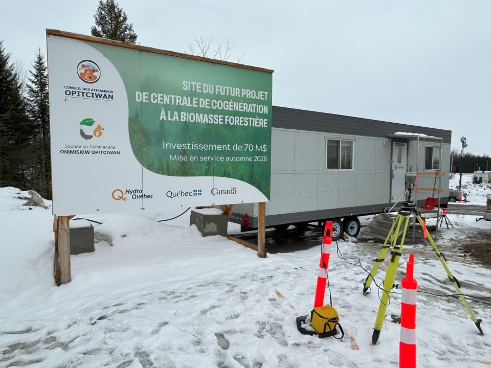 Un panneau annonçant la future centrale de cogénération à la biomasse forestière à Opitciwan est installé à côté d'une remorque de chantier temporaire dans un paysage enneigé.