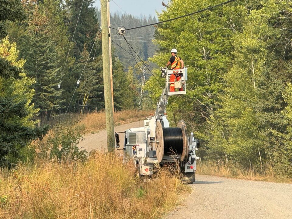 Worker in a high‑visibility safety vest and hard hat stands on an elevated bucket attached to a utility truck, installing broadband infrastructure on overhead lines at a roadside utility pole. The truck is parked on a narrow rural road with tall dry grass and dense green trees on both sides. A large spool of cable is mounted on the truck, with forested hills in the background.