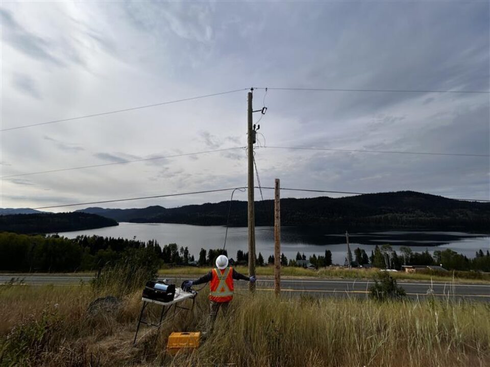 Person in a high-visibility safety vest and hard hat installs broadband infrastructure beside a roadside utility pole, handling cabling connected to overhead lines. Equipment is set up in tall grass near a two‑lane road, with a lake, forested hills, and cloudy sky in the background.