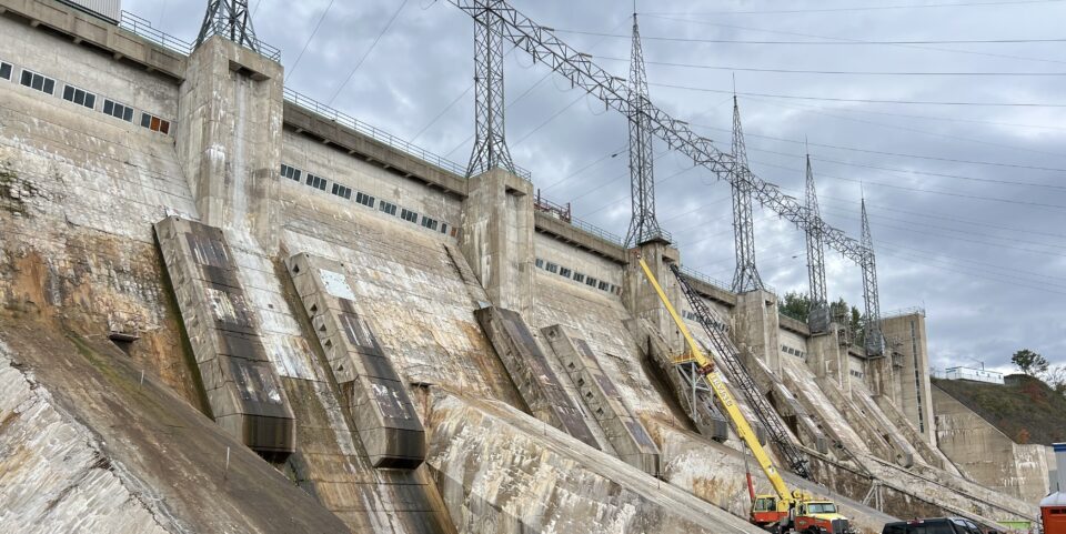 Close-up of the Mactaquac Generating Station showing the concrete dam structure, transmission towers, and maintenance equipment during rehabilitation work.