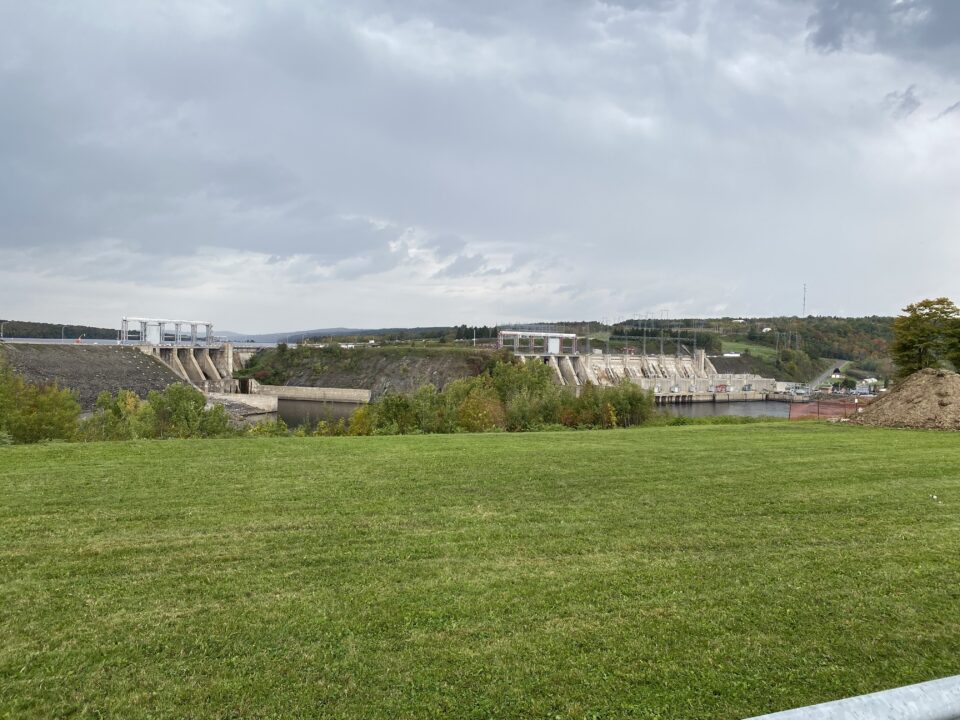 Wide view of the Mactaquac Generating Station on the Saint John River, with dam infrastructure, green lawn in the foreground, and dark storm clouds overhead.