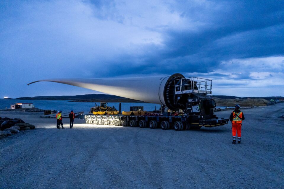Industrial construction site in a rocky Arctic landscape, with cranes, buildings and equipment beneath a cliff. In the foreground, a wind turbine blade is transported along a gravel road under an overcast sky.