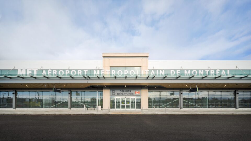 Vue frontale de l’entrée de l’aérogare pour vols intérieurs de l’aéroport métropolitain de Montréal (YHU), avec sa façade en verre, ses portes d’entrée coulissantes et le nom du bâtiment inscrit sur l’auvent extérieur supérieur.