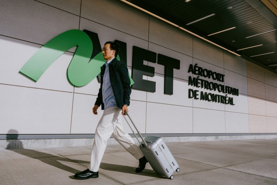 Une personne marchant à l’extérieur de l’aérogare de l’aéroport métropolitain de Montréal (YHU), tirant une valise à roulettes, avec le nom de l’aéroport « Aéroport métropolitain de Montréal » affiché sur le mur extérieur, à côté d’un grand logo MET vert.