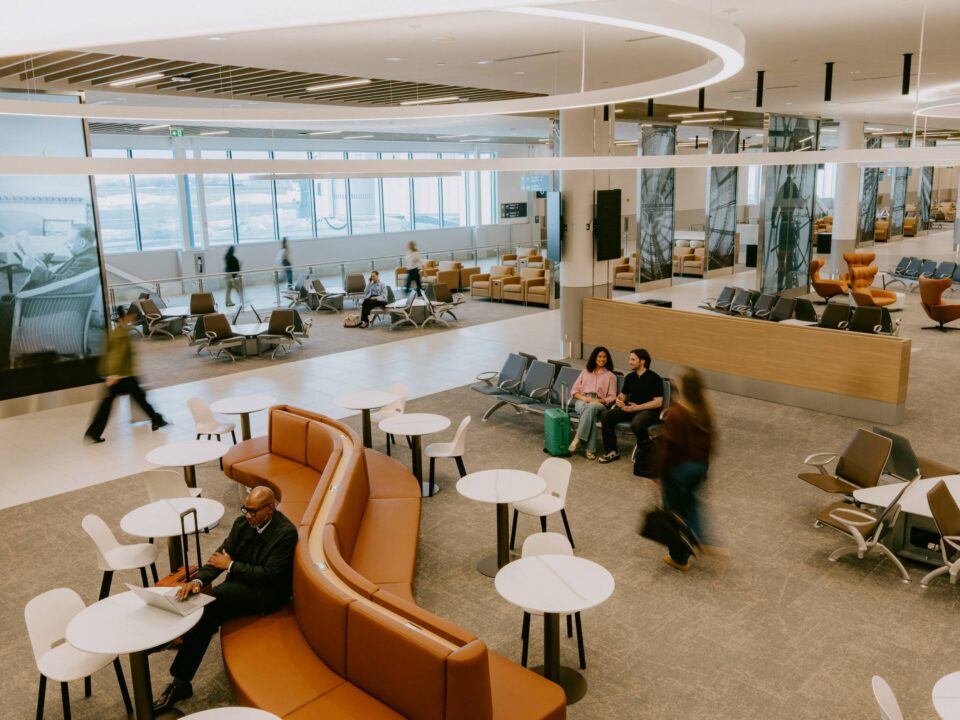 Vue intérieure de l’aérogare pour vols nationaux de l’aéroport métropolitain de Montréal (YHU), montrant un vaste hall des départs doté de rangées de sièges, de banquettes incurvées, de petites tables rondes et de grandes baies vitrées le long du mur extérieur. Plusieurs passager·ères sont assis·es ou se promènent dans l’espace, où l’on remarque partout des luminaires modernes et des finitions architecturales épurées.