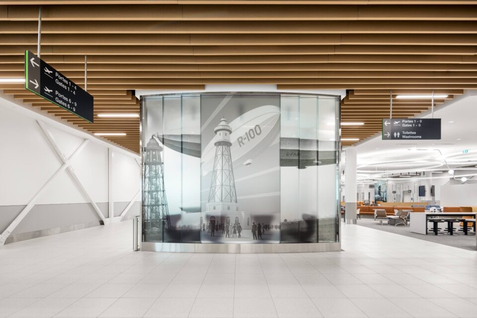 Vue intérieure du hall de l’aérogare de l’aéroport métropolitain de Montréal (YHU), où se trouve une installation circulaire en verre représentant une image d’archive d’un avion et d’une tour de contrôle. Des panneaux de signalisation suspendus guident les passager·ères vers les portes d’embarquement et les services, tandis que l’espace se caractérise par un sol blanc, des éléments de plafond lambrissés et des zones de repos visibles à l’arrière-plan.