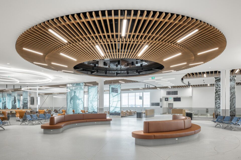 Vue intérieure de l’aérogare de l’aéroport métropolitain de Montréal (YHU) montrant un hall ouvert doté de banquettes incurvées et d’un grand plafond circulaire en bois avec éclairage intégré. L’espace comprend de larges allées, des zones de repos disséminées et des cloisons en verre ornées de panneaux illustrés, créant ainsi un environnement moderne, lumineux et spacieux.