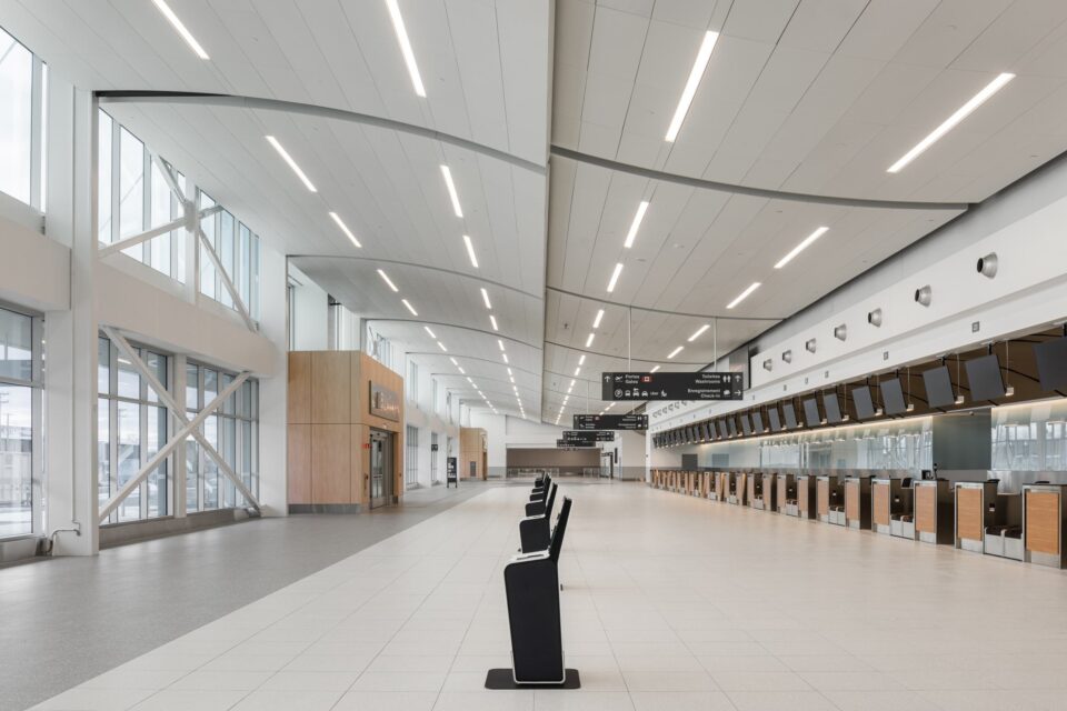 Vue intérieure de la zone d’enregistrement de l’aérogare de l’aéroport métropolitain de Montréal (YHU), montrant une longue rangée de comptoirs d’enregistrement surmontés d’enseignes, un vaste hall ouvert avec de hauts plafonds et un éclairage linéaire, ainsi qu’un petit nombre de bornes autonomes disposées au centre de l’espace. De grandes baies vitrées bordent le côté gauche, laissant pénétrer la lumière naturelle dans ce hall épuré et moderne.