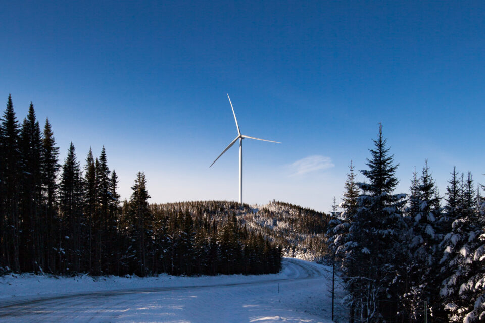 Snowy landscape with a large wind turbine atop a hill, surrounded by evergreen trees.