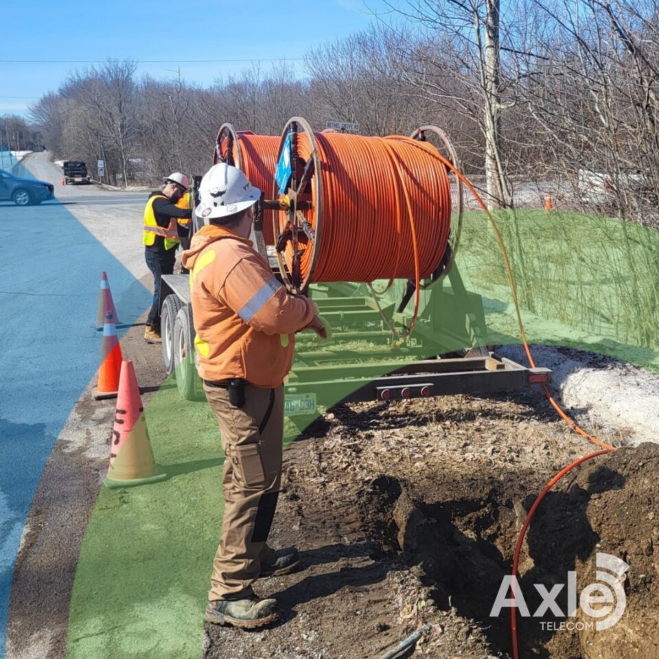 Workers in orange safety vests near a large orange cable reel, digging a trench for fiber optic cable installation.