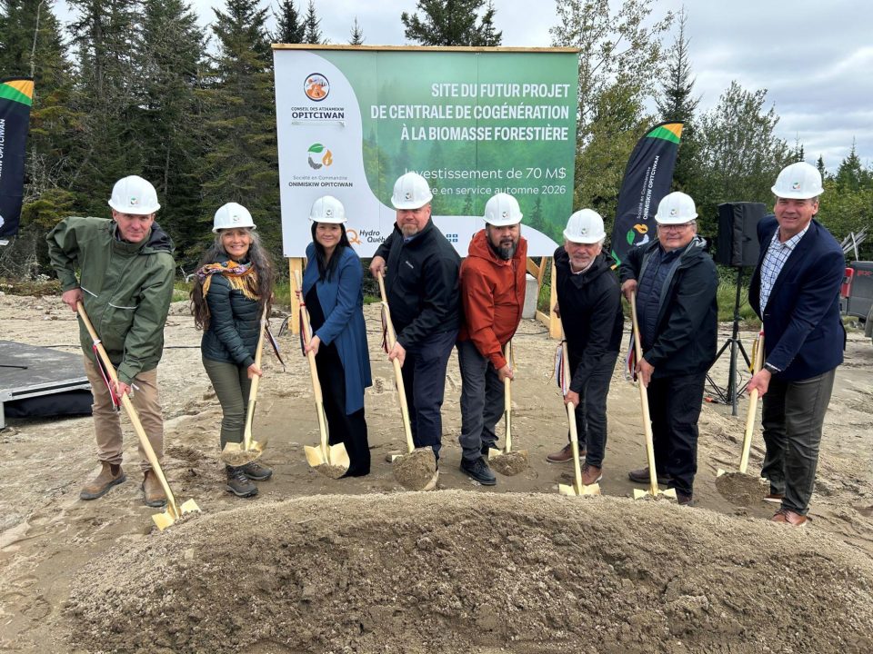 Un groupe de huit personnes avec des casques de chantier participe à la cérémonie d'inauguration du début des travaux des futures installations de la centrale de cogénération à la biomasse forestière à Opitciwan, tenant des pelles devant un grand panneau.