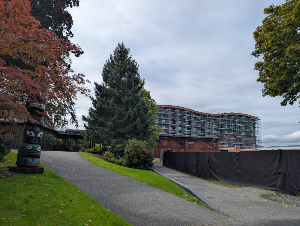 Landscaped pathway leading toward a large building under construction, with scaffolding covering the exterior. Trees with autumn foliage and a carved totem pole are visible in the foreground.