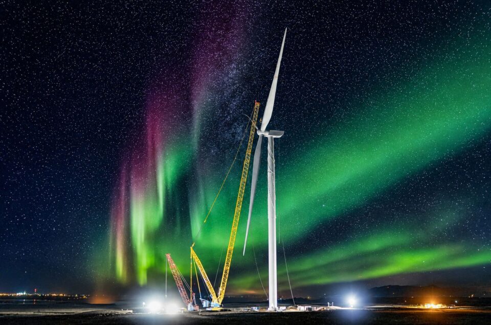 Wind turbine under construction at night, illuminated by cranes and lights, with the northern lights visible in the sky above.