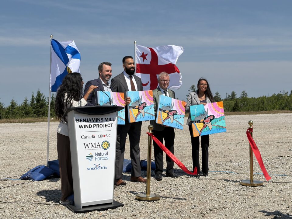 Plusieurs personnes se tiennent à l'extérieur lors d'une cérémonie d'inauguration du projet d'énergie éolienne de Benjamins Mill. Un podium au premier plan affiche les logos du projet et des partenaires, tandis que les drapeaux de la Nouvelle-Écosse et mi'kmaq sont hissés derrière le groupe. Les participant·e·s tiennent des œuvres d'art colorées et se tiennent près d'un ruban rouge sur un site de gravier sous un ciel bleu dégagé. Le texte visible sur le podium comprend « projet d'énergie éolienne de Benjamins Mill » ainsi que les logos de Natural Forces, de WMA, de la BIC et du gouvernement du Canada.