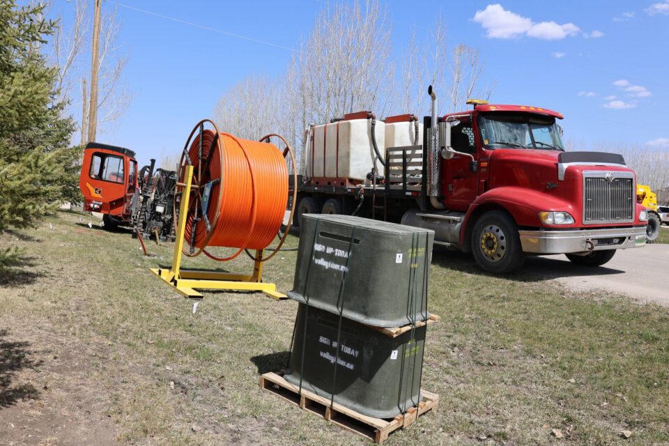 Bright day scene with a red truck, broadband construction equipment, including a fibre cable spool and service boxes, along a rural roadside. Leafless trees and a blue sky are in the background.