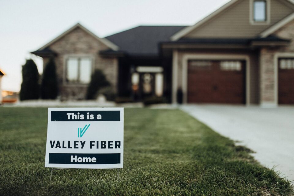 A sign on a lush green lawn reads “This is a Valley Fiber Home,” in front of a large, modern brick house with a driveway and garage.