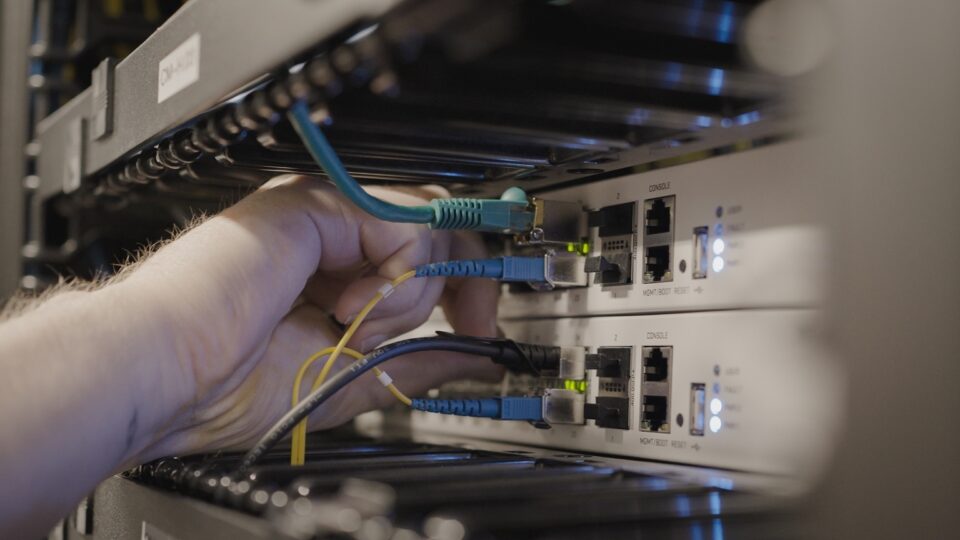 Une main connectant des câbles réseau bleus et jaunes à des commutateurs à l’intérieur d’une armoire de serveurs, dont les voyants lumineux sont allumés
