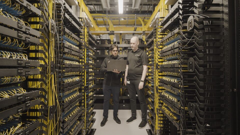 Deux techniciens se tiennent entre des rangées de baies de serveurs dans un centre de données, en train de consulter des informations sur un ordinateur portable au milieu de câbles jaunes soigneusement rangés.