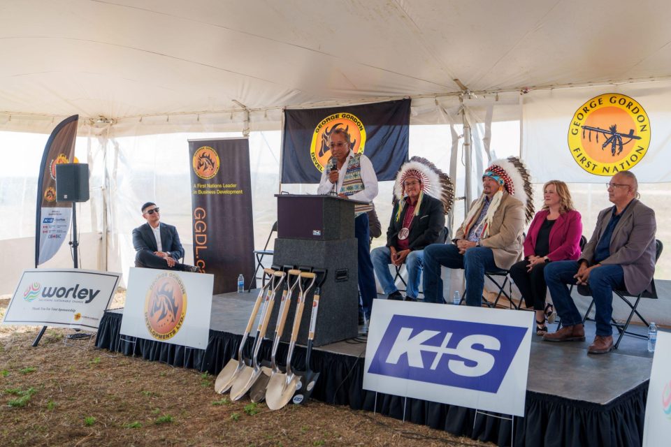 Le chef de la PNGG, portant une coiffe à plumes et une veste beige, parle du partenariat avec Wicehtowak depuis une tribune installée sous une tente, entouré d’autres personnes assises et de pelles cérémonielles disposées devant lui. Des bannières arborant le logo « George Gordon Developments Ltd. » sont affichées derrière lui.