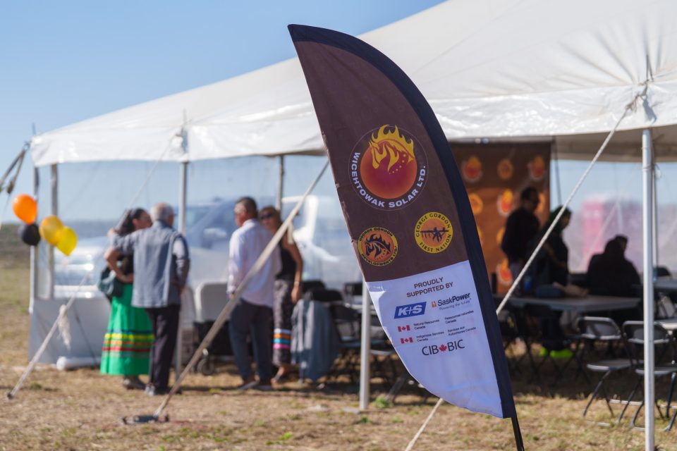 Un drapeau promotionnel de Wicehtowak Solar Ltd. se dresse au premier plan devant une tente blanche lors de l’événement annonçant le partenariat, où des personnes se sont rassemblées par une journée ensoleillée.