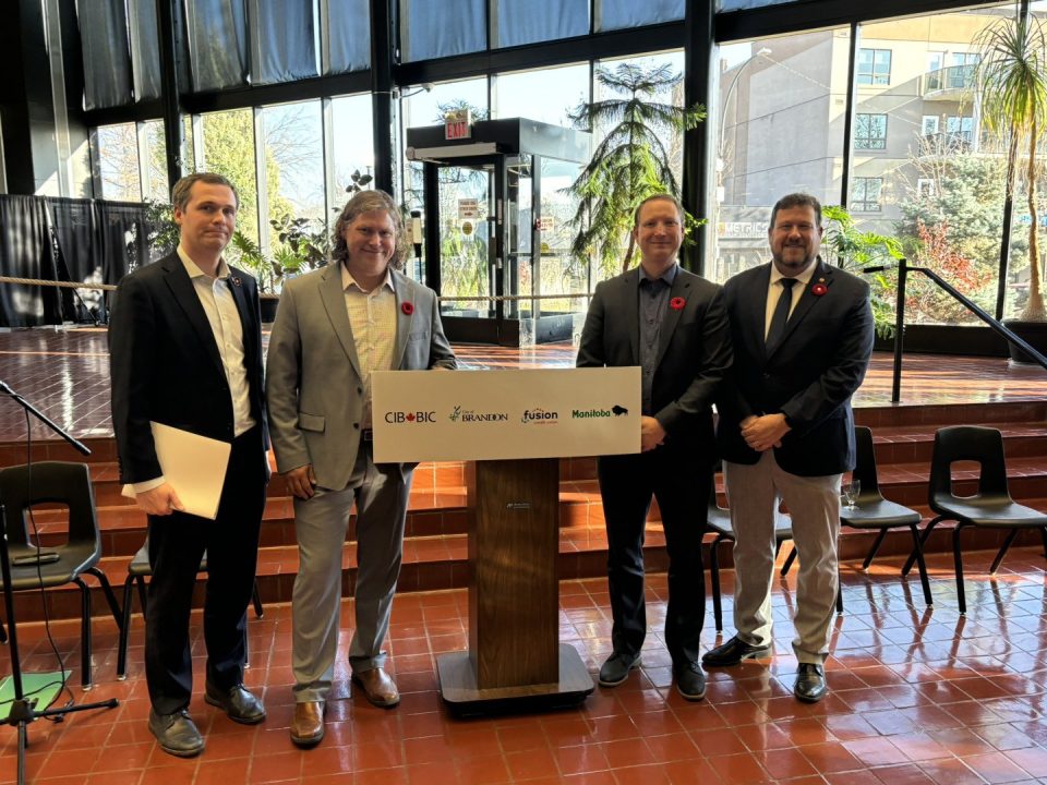 Quatre hommes en costume, Alex Ryan (BIC), Jeff Fawcett (maire de Brandon), Darwin Johns (PDG, Fusion Credit Union) et Glen Simard (député provincial, Brandon-Est), se tiennent derrière un podium en bois arborant les logos de la BIC, de Fusion Credit Union et de la province du Manitoba, dans une grande salle aux larges fenêtres donnant sur la végétation, lors de la conférence de presse sur le projet d’infrastructures liées à l’eau de Brandon, le 5 novembre.