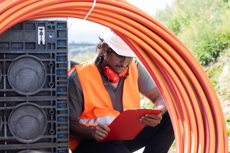 A worker in an orange safety vest and white hard hat, wearing red earmuffs, sits reviewing documents on a clipboard while surrounded by coiled orange fibre cables.