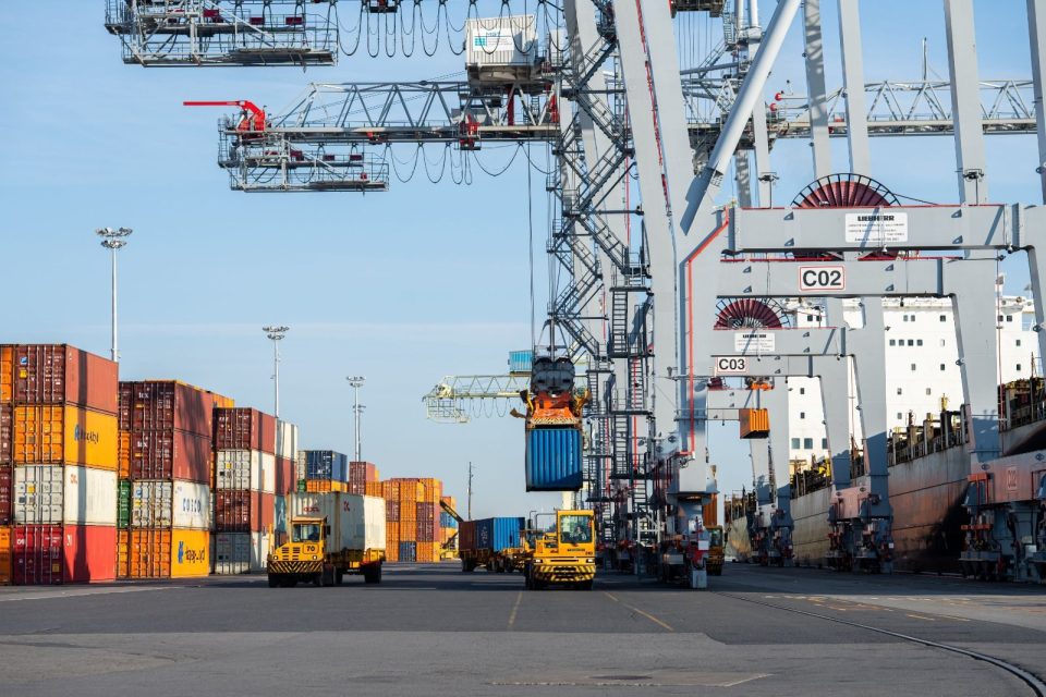 Large cranes load containers onto a ship at a terminal while other metal containers sit on the dock.