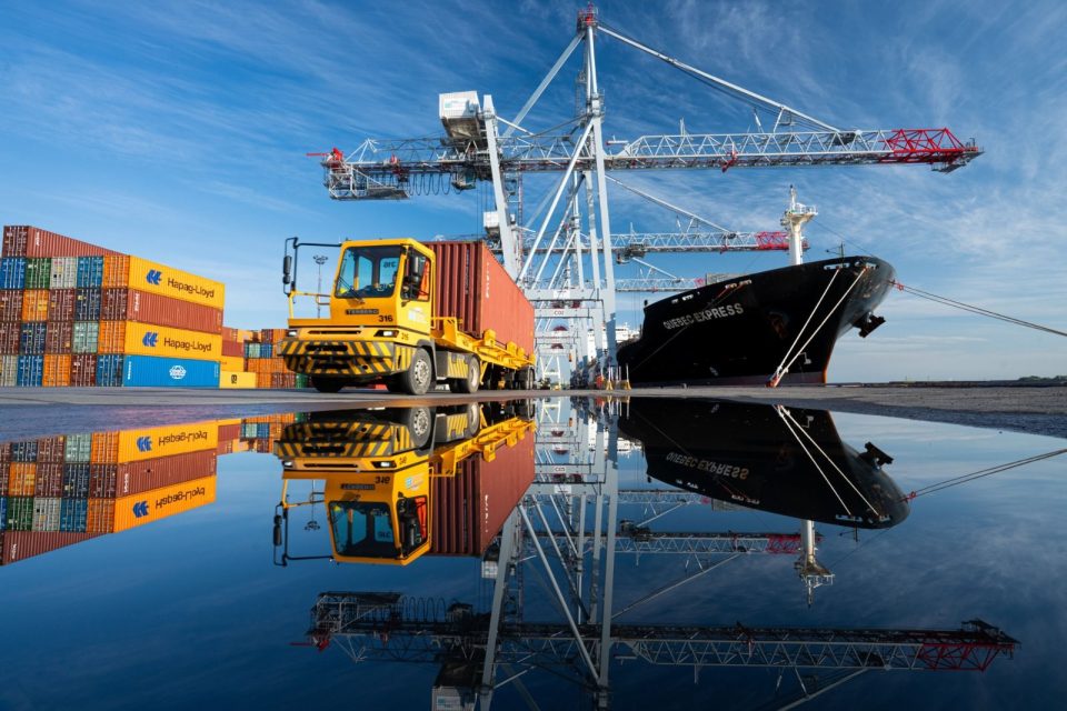 Containers sit at a dock and on a truck near cranes used to load a ship that sits under blue skies.