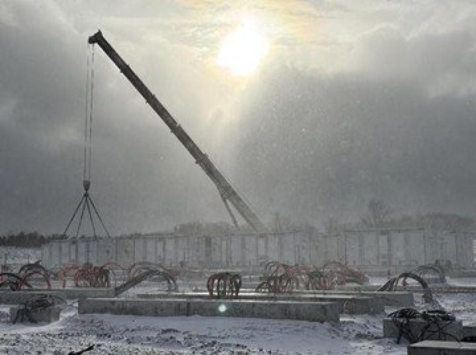 Construction hivernale du site de stockage de batteries de White Rock avec une grande grue soulevant des matériaux sous la neige qui tombe, éclairant les coffrages en béton au sol.