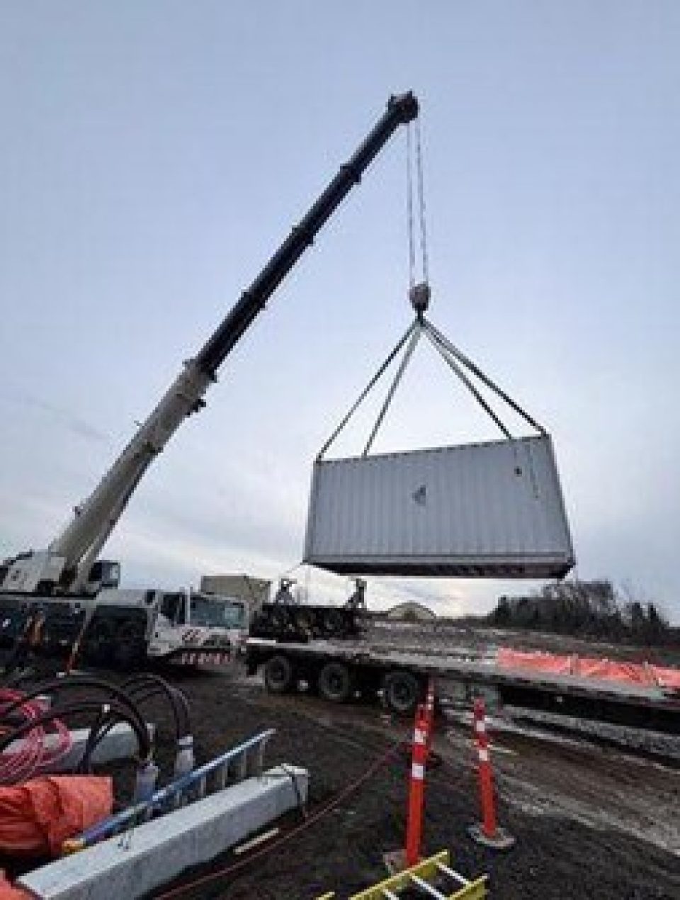Une grande grue mobile soulève un conteneur d’expédition blanc au-dessus d’une remorque à plateau pendant la construction hivernale du site de stockage de batteries de White Rock.